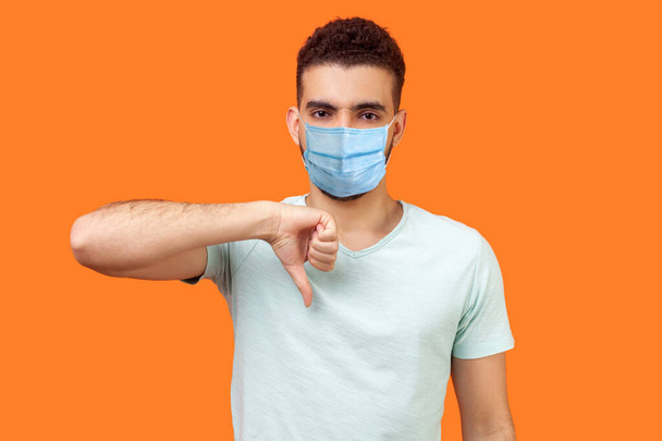 Thumbs down! Portrait of frustrated dissatisfied man with medical mask in casual white t-shirt showing dislike gesture, disapproval negative sign. indoor studio shot isolated on orange background - Фото, зображення