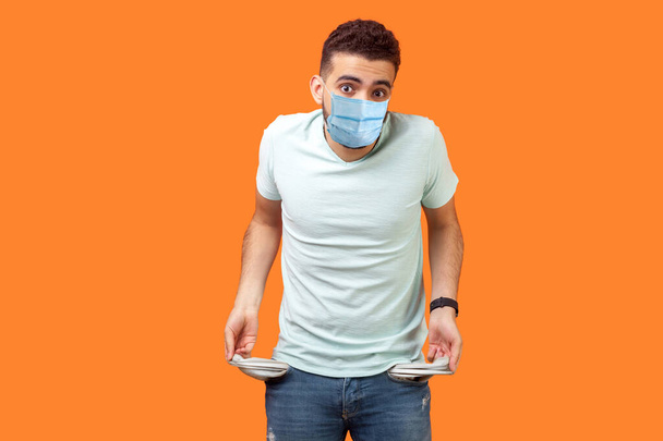 Portrait of frustrated worried man with medical mask in casual white t-shirt turning out empty pockets showing I have no money gesture, bankrupt. indoor studio shot isolated on orange background - Foto, immagini