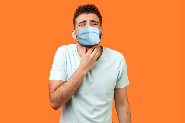 Inflamed throat. Portrait of sick man with medical mask in white t-shirt touching painful neck, suffering sore throat, tonsillitis symptom, medical concept. studio shot isolated on orange background - Photo, image