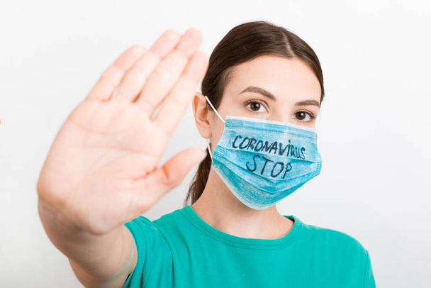 Πορτρέτο μιας γυναίκας με ιατρική μάσκα με κείμενο stop coronavirus σε λευκό φόντο. Το άτομο δείχνει χειρονομία στάσης. Ιός του κερατοειδούς. Αναπνευστική προστασία. - Φωτογραφία, εικόνα