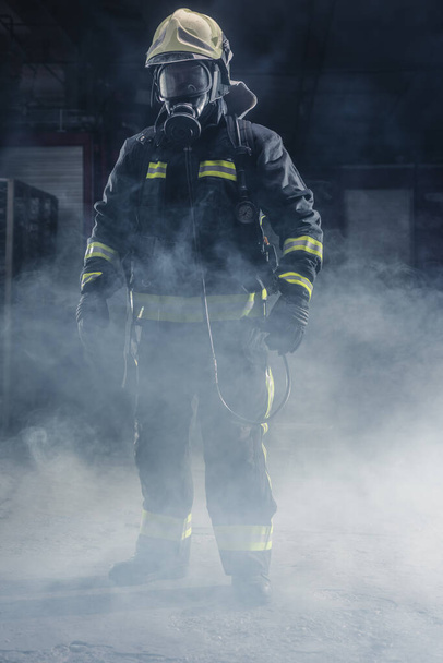 Portret strażaka noszącego frekwencję strażacką i kask. Ciemne tło z dymem i niebieskim światłem. - Zdjęcie, obraz