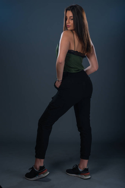 Portrait of a sexy young female model  standing in a studio and wearing dark green top and black pants - Fotó, kép