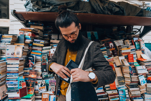 Moderno jovem hipster segurando um saco e um livro enquanto caminhava pela praça da cidade
 - Foto, Imagem