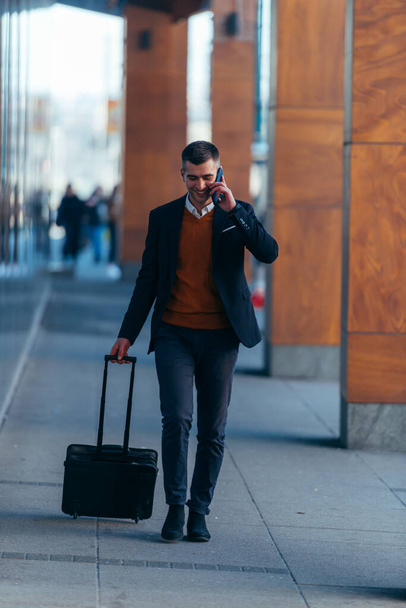 Young businessman walking through a station while talking on the phone and pulling his travel bag. - Foto, imagen