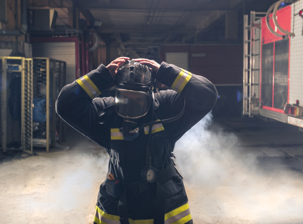 Portré egy tűzoltóról, aki tűzoltó felszerelést és sisakot visel. Sötét háttér füst és kék fény. - Fotó, kép