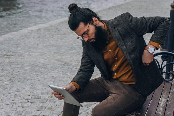 Hipster-Mann in modernem Outfit sitzt auf einer Bank und checkt seine E-Mails auf einem Tablet - Foto, Bild