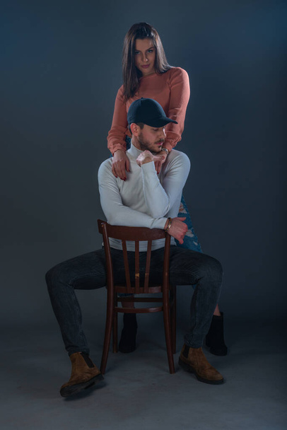 Красивий чоловік сидить на стільці і красива жінка, що стоїть поруч з ним на темному тлі в студії
 - Фото, зображення