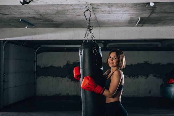 Imagen de una joven sonriente y fuerte sosteniendo una bolsa de boxeo dentro del garaje
 - Foto, Imagen