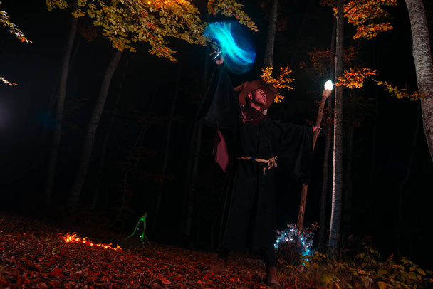 Hechicero (mago) lanzando un hechizo mientras está de pie en los bosques mágicos. Sostiene su bastón mágico. La iluminación mágica. Celebración de vacaciones de otoño. Concepto de misterio y pesadilla - Foto, imagen