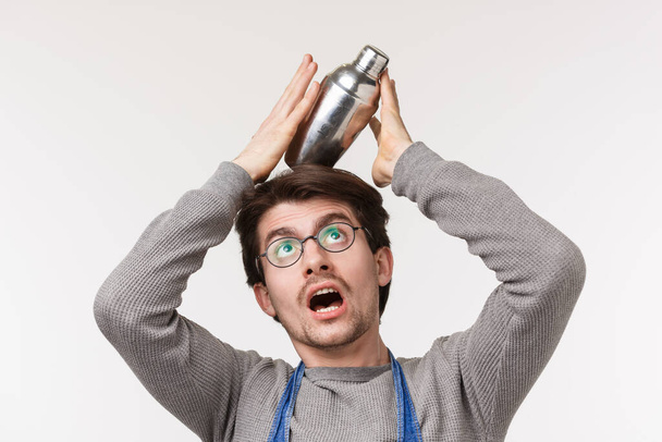 Close-up portrait of funny caucasian male bartender in apron and glasses balancing shaker on head as its falling on ground, standing white background fool around while there are no customers - Photo, Image