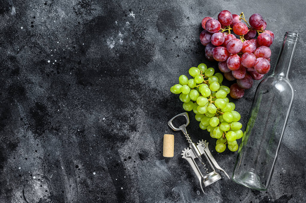 Een tak van groene en rode druiven, een fles, een kurkentrekker en een kurk. Concept van de wijnbereiding. Zwarte achtergrond. Bovenaanzicht. Kopieerruimte. - Foto, afbeelding