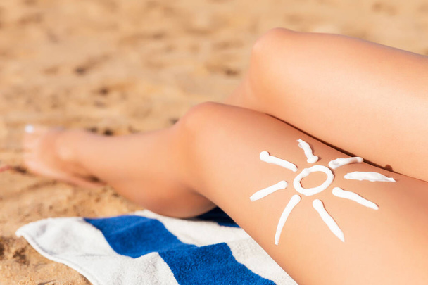 Zonnebrandcrème wordt gemaakt in de vorm van de zon op een gebruinde vrouwen poot die ontspant op de handdoek op het strand. Kanker zorgconcept. - Foto, afbeelding