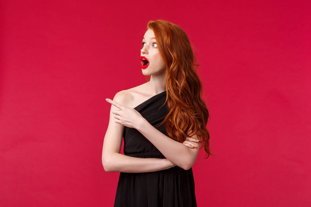 Celebration, events, fashion concept. Portrait of amazed and shocked redhead young pretty woman in elegant black evening dress, drop jaw, looking and pointing left astonished, red background - 写真・画像