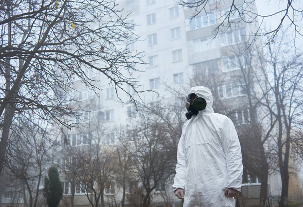 Side view of ecologist in protective uniform standing on foggy street and looking aside. Male environmentalist wearing white suit and gas mask. Concept of ecology and environmental pollution. - Fotografie, Obrázek