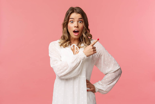 Surprised beautiful blond girl pointing upper right corner, gasping amused and staring at camera with fascinated expression, open mouth in awe to ask question about promo, pink background - Photo, Image