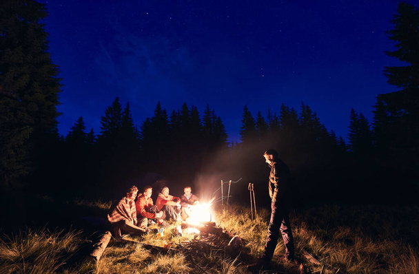 Friends hikers having a rest at evening by burning fire at campsite in forest. Tourists people are sitting on log against the backdrop of mighty fir trees and night sky on which stars appear - Φωτογραφία, εικόνα