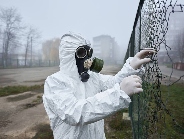 Zbliżenie męskiego naukowca rozrywającego drut ogrodzeniowy. Mężczyzna w masce gazowej i kostium ochronny stojący na pustym boisku do piłki nożnej w pobliżu budynku - Zdjęcie, obraz