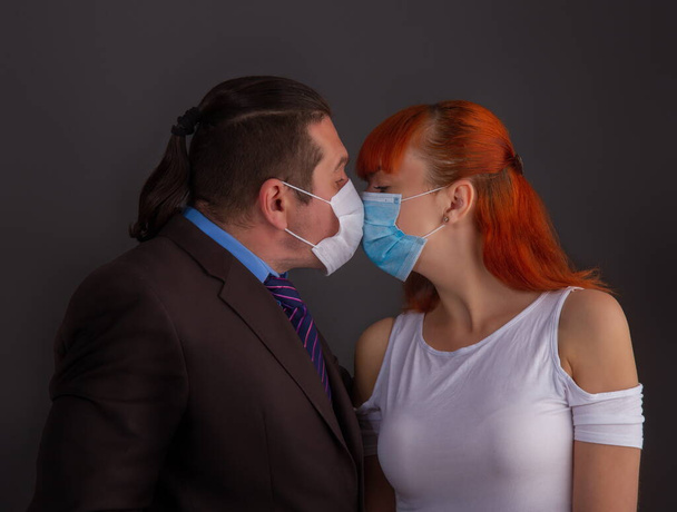 A man in a suit and a girl kiss on a dark background through protective medical masks. - Valokuva, kuva