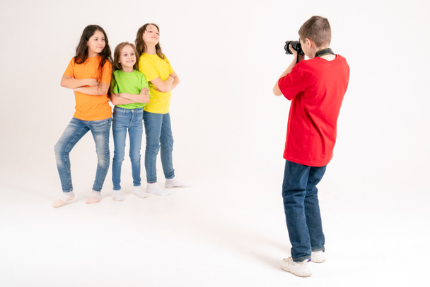 Chłopiec fotograf robi zdjęcia trzem uroczym dziewczynom w jasnych koszulkach na białym tle w studio. Młody fotograf i bloger. - Zdjęcie, obraz
