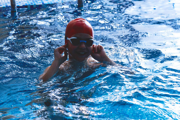 Joyful smiling boy swimmer in a cap and Goggles learns professional swimming in the swimming pool in gym close up - Photo, Image