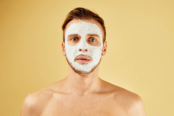 Portrait of a bearded puzzled man with open mouth and white mask on his face in the studio isolated on a yellow background, skin care concept - Фото, изображение