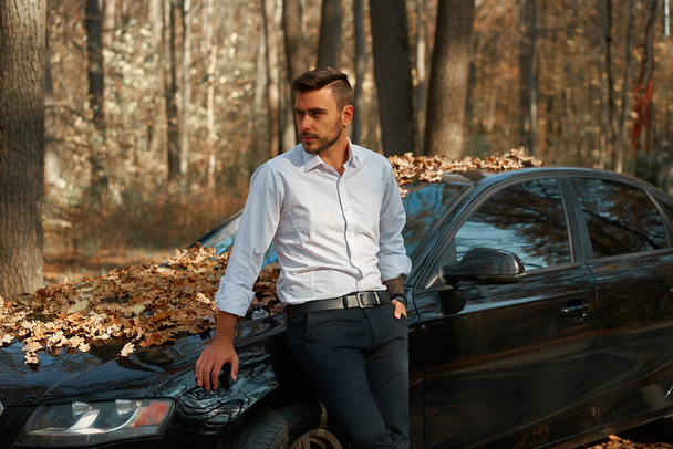A handsome Caucasian man wearing business suit standing near black car on nature.  Young adult businessman travels by car on a beautiful sunny autumn day. Elite luxury driver. - Fotoğraf, Görsel