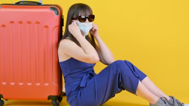 A young girl in sunglasses sits near a suitcase and takes off a medical mask. Torn vacation. Isolated on a yellow background. ealth care and medical concept. - Video