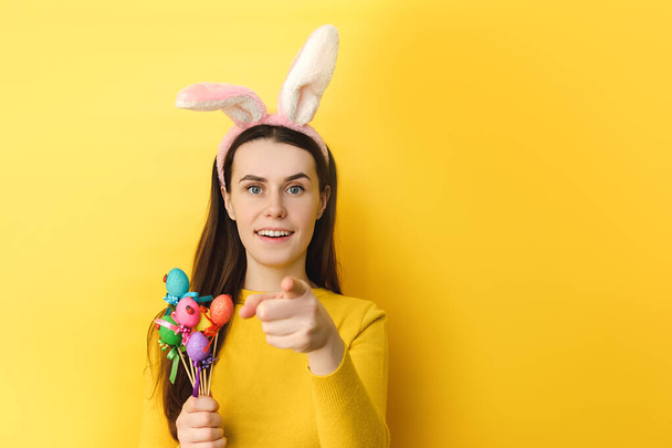 Positive woman with bunny fluffy ears, has happy surprised look, wears bunny ears on head, looks at camera, holds painted eggs on sticks, stands over yellow studio wall. Seasonal holiday concept. - Фото, зображення