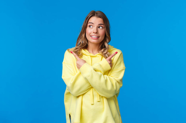 Various choices, cant decide. Cute dreamy blond girl in yellow hoodie making her choice, pointing fingers sideways at left and right side and smiling, picking one thing from all, blue background - Photo, Image