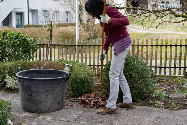Junge Frau harkt Blätter im Frühling. Fußweg und Blumenbeet im Vorgarten reinigen. Ländliche Szene. - Foto, Bild