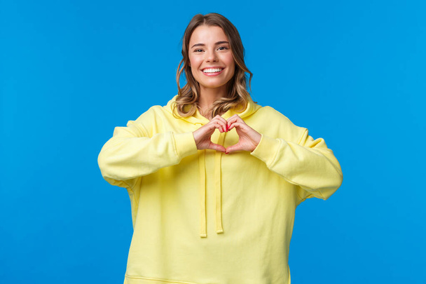Heerlijk verzorgend schattig blond meisje in gele hoodie tonen hart gebaar en glimlachen als uitdrukkelijke sympathie, bekennen in liefde of hartverwarmende gevoelens naar persoon, staan blauwe achtergrond - Foto, afbeelding