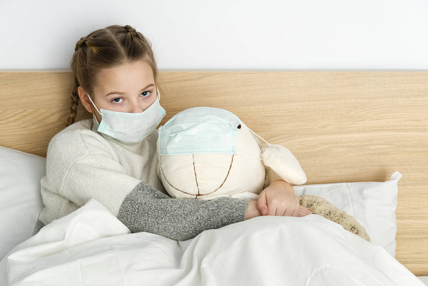 Ateşi ve baş ağrısı olan maskeli hasta üzgün bir çocuk yatakta yatıyor. Grip salgını virüsü bakterisi. Yatak odasında oyuncak taşıyan bir kız koruyucu maske takıyor, Coronavirüs konseptine karşı koruyor.. - Fotoğraf, Görsel