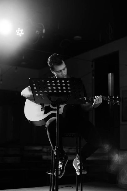 Mladý pohledný muž evropského vzhledu hraje na akustickou kytaru a sedí na židli. Černobílá fotografie. - Fotografie, Obrázek
