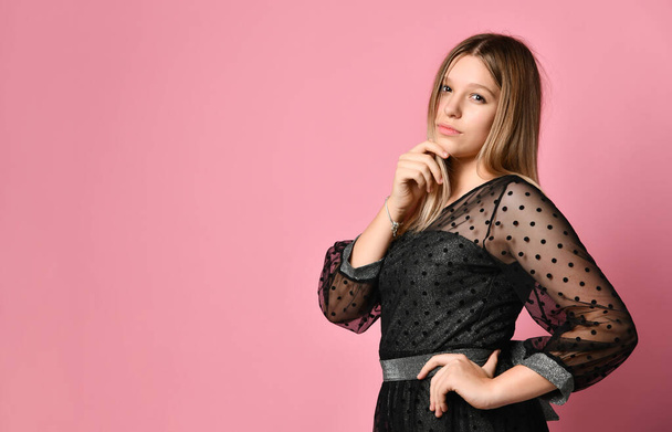 Teenage female in black dress. She is looking at you, touching her chin, posing sideways against pink studio background. Close up - Foto, immagini