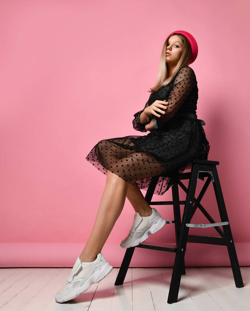 Teen girl in black dress, red hat and white sneakers. She sitting on step ladder, posing sideways on pink background. Full length - Foto, immagini