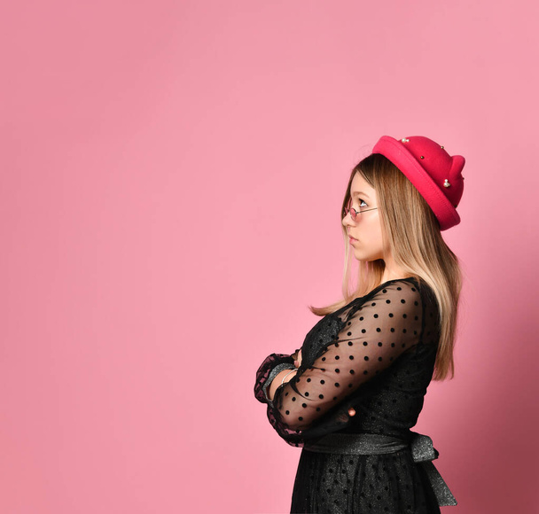 Teenager in sunglasses, black dress and red hat. She is looking up, hands folded, posing sideways on pink background. Close up - Foto, Imagen