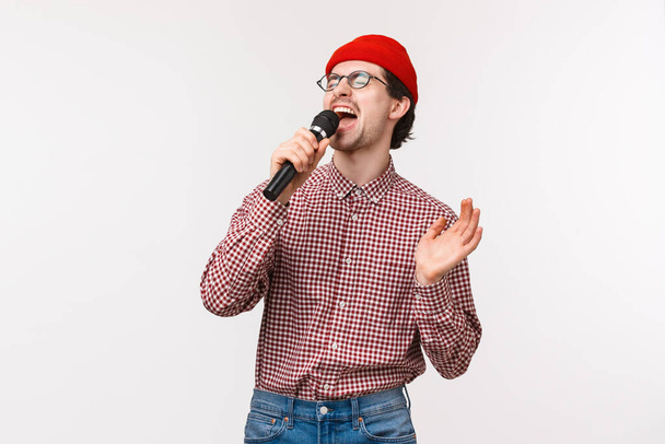 Brokenhearted guy sing romantisches Lied in Karaoke mit leidenschaftlichem Gefühl, Augen zu und schreien ins Mikrofon, steht in roter Mütze, Brille und kariertem Hemd, weißer Hintergrund - Foto, Bild