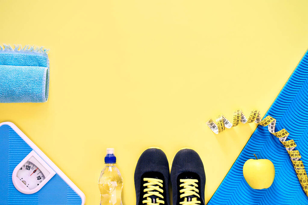 Sportschuhe, Wasserflasche, Apfel, Waage zum Wiegen und Maßband auf gelbem Hintergrund. Sportgeräte. Gesunder Lebensstil und Ernährungskonzept. Gelb und hellblau. Kopierraum. Ansicht von oben. - Foto, Bild