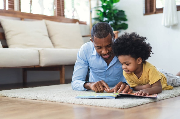 Happy African American father and son are reading a book and smiling while lying on floor spending time together at home. Children education and development concept. - 写真・画像