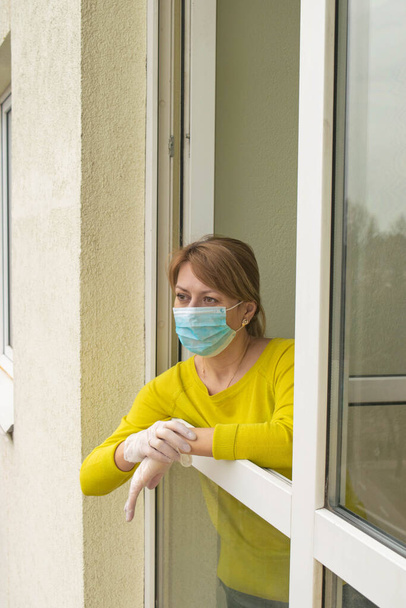 Die Frau mit Schutzmaske und Handschuhen steht am offenen Fenster ihrer Wohnung und folgt den Empfehlungen der Selbstisolierung und des Heimbleibens während der Epidemie und Quarantäne.. - Foto, Bild