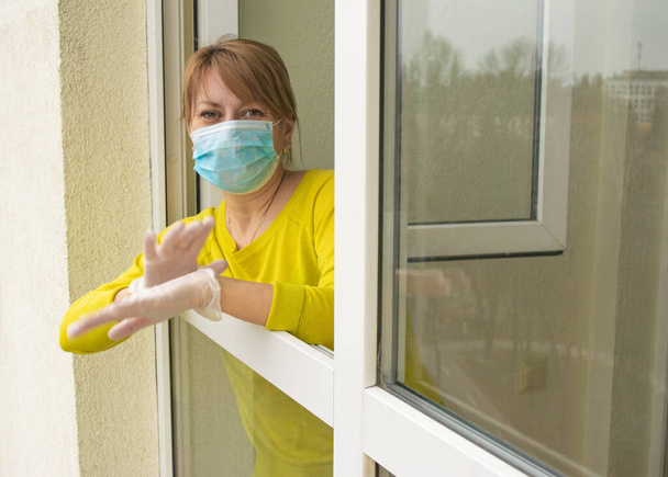 Die Frau mit Schutzmaske und Handschuhen steht am offenen Fenster ihrer Wohnung und folgt den Empfehlungen der Selbstisolierung und des Heimbleibens während der Epidemie und Quarantäne.. - Foto, Bild