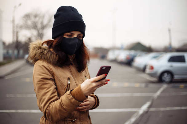 Young girl in city street wearing black sterile medical face mask. Woman using the phone to search for news about nCov 2019. Quarantine COVID-19 pandemic coronavirus epidemic and health care concept - Foto, afbeelding