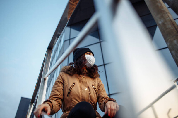 Een jong meisje met een medisch masker zit op een trap van een gesloten winkelcentrum. Bescherming van de luchtwegen. Coronavirus. Gevaarlijke ziekte. Artikel over coronavirus en de behandeling ervan - Foto, afbeelding