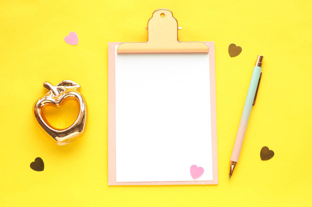 a board with a clip with a white sheet mock-up, a golden apple figurine with a hole in the heart, a pink-blue pen, candy hearts on a bright yellow background. flat layout, top view. - Photo, Image