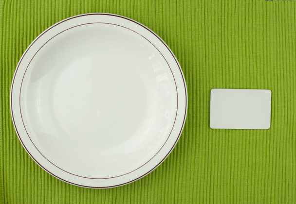 Vista superior. Placa de cerâmica com etiqueta vazia na toalha de mesa verde
 - Foto, Imagem