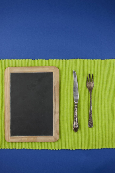 marco de pizarra de menú con cuchillo y tenedor para recetas o menú sobre fondo azul con mantel verde
 - Foto, Imagen