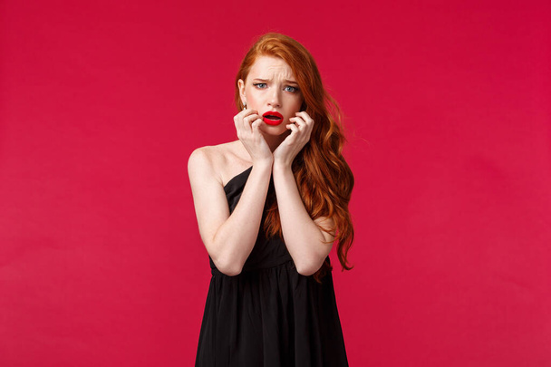 Fashion, luxury and beauty concept. Portrait of timid and scared ginger girl in black dress feel unsafe or insecure, touch face frowning frightened, look concerned and worried camera, red background - Photo, Image