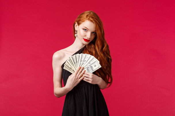 Luxury, beauty and money concept. Portrait of sensual good-looking, flirty young redhead woman in black stylish dress, hinting you can get this cash dollars if place good bet, smiling daring - Foto, Imagem