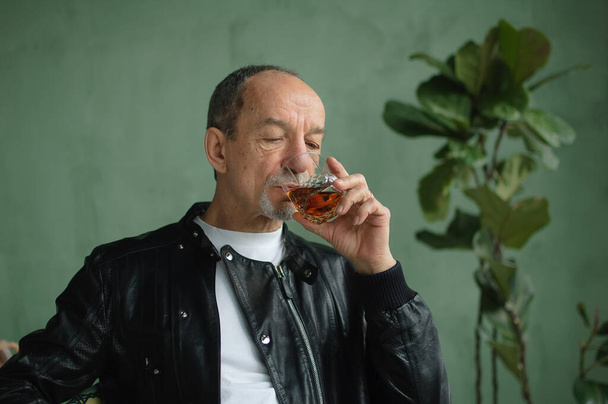 Senior άνθρωπος σε μαύρο δερμάτινο μπουφάν πίνει ουίσκι από γυαλί σε σοφίτα στυλ δωμάτιο με ανοιχτό πράσινο τοίχους και φυτά στο παρασκήνιο. Εξάρτηση αλκοόλ - Φωτογραφία, εικόνα