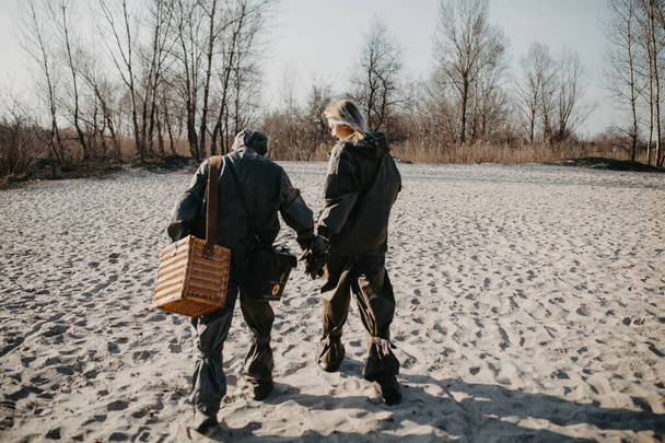 Couple in love walks at beach in NBC protective suits. Concept of a preventive measures and protection for coronavirus COVID 19 pandemic and other global dangers. - Photo, Image